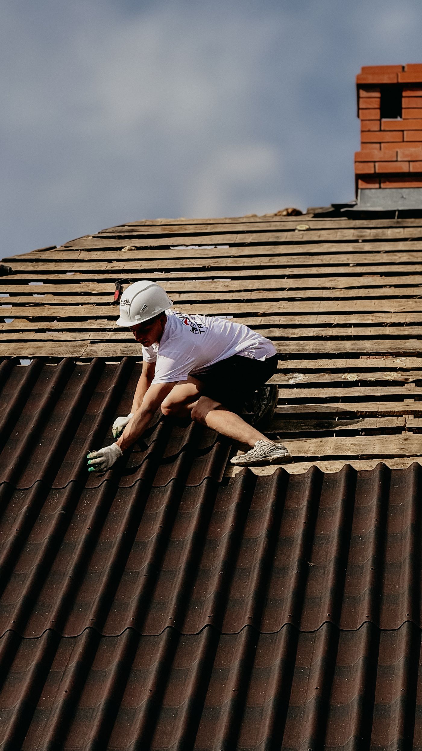 Dachdecker mit Helm arbeitet an der Eindeckung eines Steildachs neben einem Backsteinkamin.